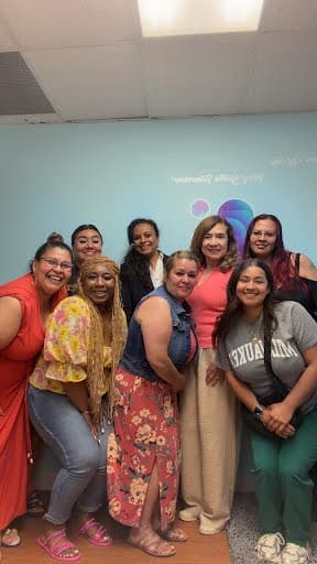 A group of women smiling together in front of the Beacon of Hope wall sign after a community gathering.